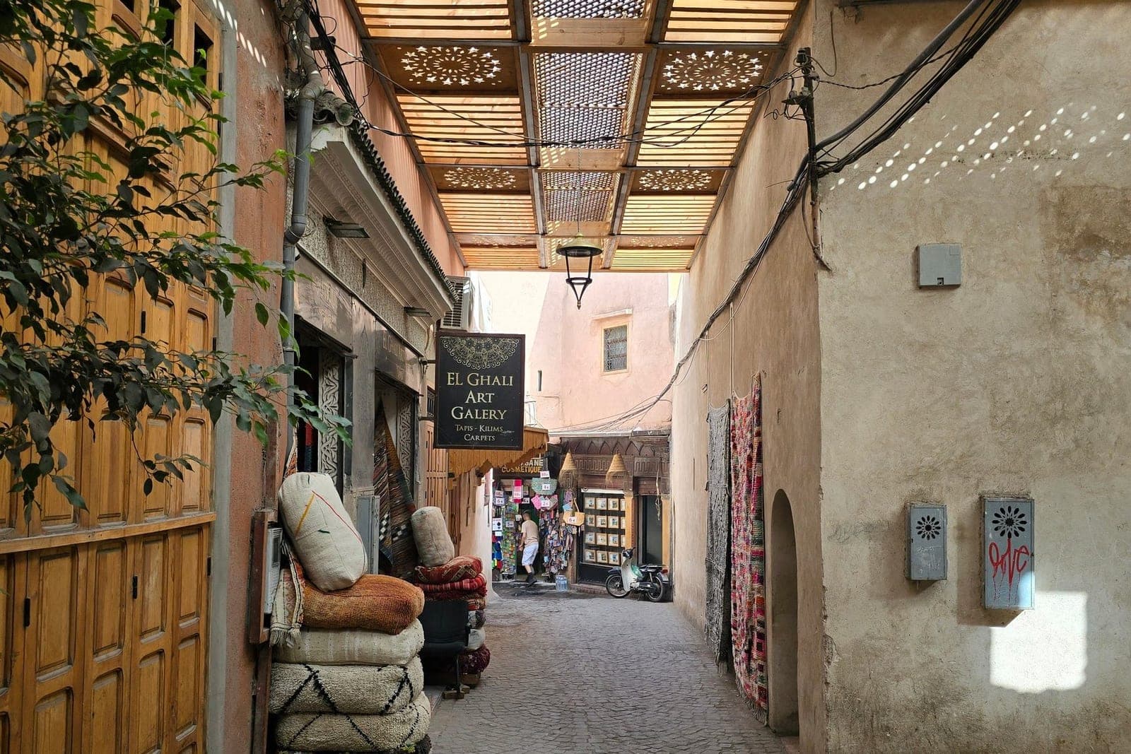 A quiet alleyway in the Marrakech medina, rugs stacked against the wall beneath a latticed roof.