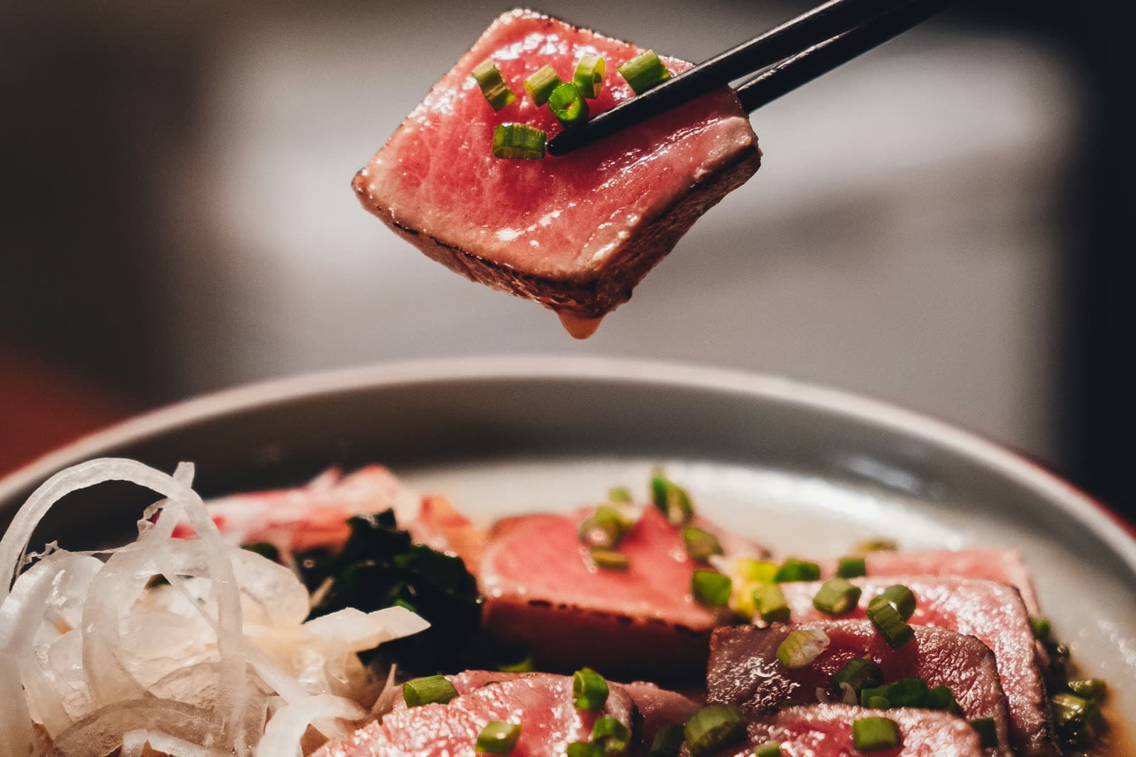 A slice of marbled wagyu held in chopsticks above a bowl of sliced beef and spring onion.