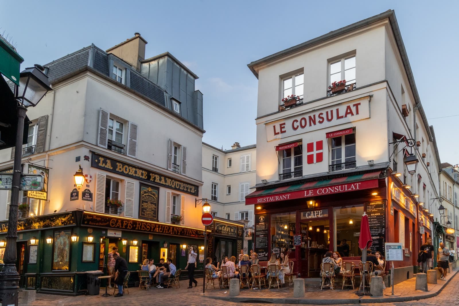 Café terraces on a Montmartre corner at dusk, Le Consulat and La Bonne Franquette lit above them.
