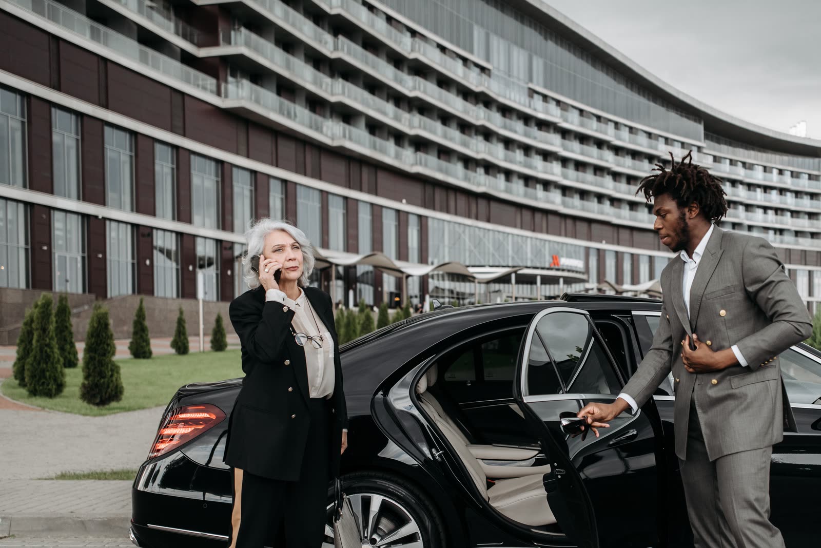 A chauffeur holds the car door open for a client outside a hotel entrance.