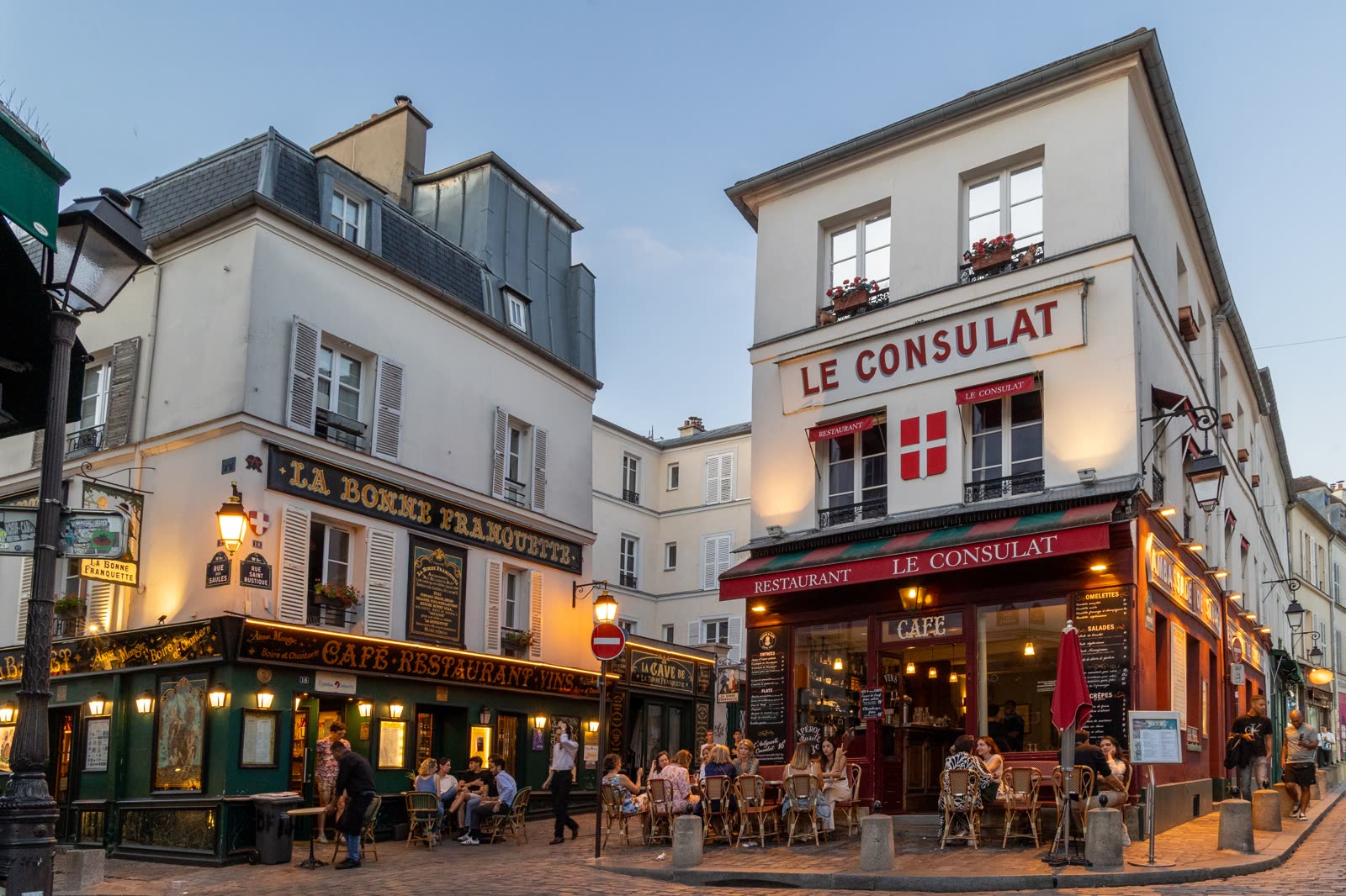 Café terraces on a Montmartre corner at dusk, Le Consulat and La Bonne Franquette lit above them.