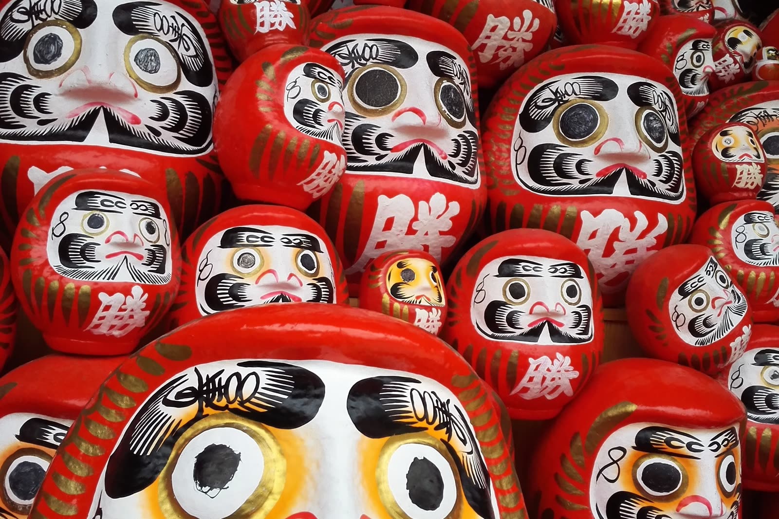 Rows of red daruma dolls stacked at a temple stall, each with a bold painted face and gold lettering.