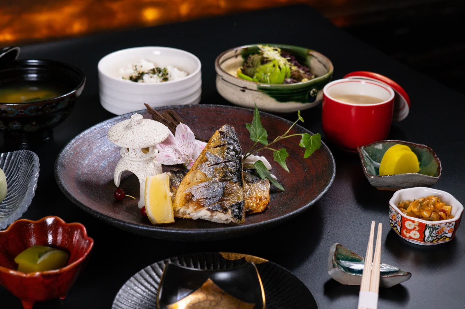 A multi-course kaiseki spread: grilled fish on dark ceramic, red lacquer bowls, rice, pickles and greens.